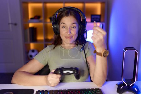 Photo for Beautiful brunette woman playing video games wearing headphones doing money gesture with hands, asking for salary payment, millionaire business - Royalty Free Image