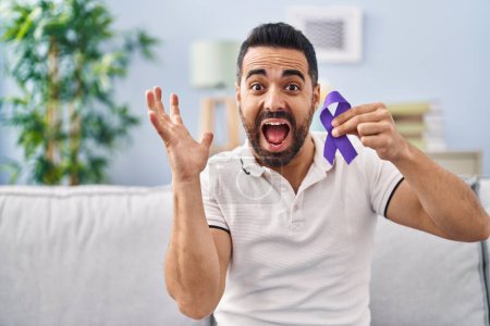 Téléchargez les photos : Jeune homme hispanique avec la barbe tenant la conscience du ruban violet célébrant la victoire avec un sourire heureux et l'expression gagnante avec les mains levées - en image libre de droit