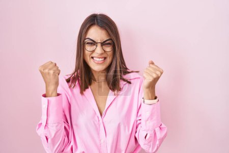 Photo for Young hispanic woman wearing glasses standing over pink background very happy and excited doing winner gesture with arms raised, smiling and screaming for success. celebration concept. - Royalty Free Image
