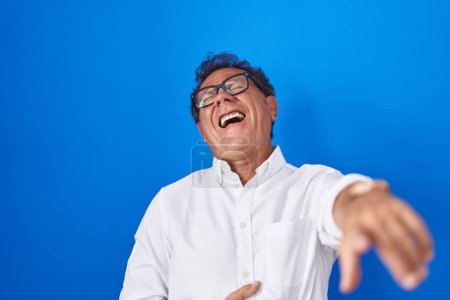 Foto de Hombre hispano de mediana edad de pie sobre fondo azul riéndose de ti, señalando con el dedo a la cámara con la mano sobre el cuerpo, expresión de vergüenza - Imagen libre de derechos