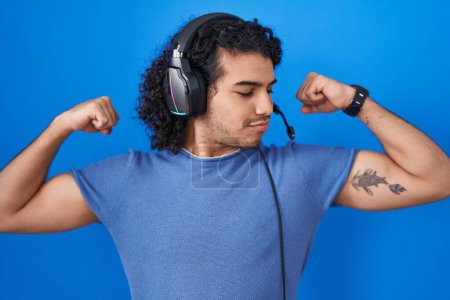 Téléchargez les photos : Homme hispanique aux cheveux bouclés écoutant de la musique à l'aide d'écouteurs montrant les muscles des bras souriant fier. concept de fitness. - en image libre de droit