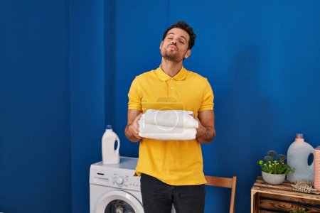 Photo for Young hispanic man holding clean towels at laundry room looking at the camera blowing a kiss being lovely and sexy. love expression. - Royalty Free Image