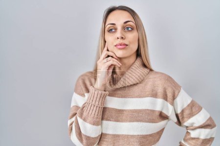 Young blonde woman wearing turtleneck sweater over isolated background with hand on chin thinking about question, pensive expression. smiling with thoughtful face. doubt concept. 