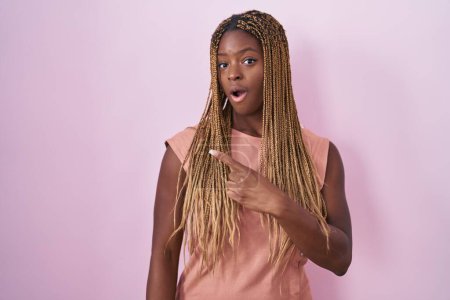 Téléchargez les photos : Femme afro-américaine avec des cheveux tressés debout sur fond rose surpris pointant du doigt sur le côté, bouche ouverte expression étonnée. - en image libre de droit