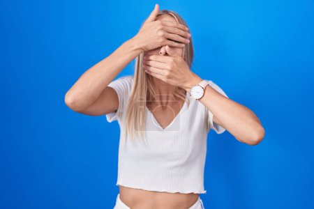 Photo for Young caucasian woman standing over blue background covering eyes and mouth with hands, surprised and shocked. hiding emotion - Royalty Free Image