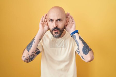 Foto de Hombre hispano con tatuajes de pie sobre fondo amarillo tratando de escuchar ambos gestos de las manos en la oreja, curioso por chismes. problema auditivo, sordo - Imagen libre de derechos