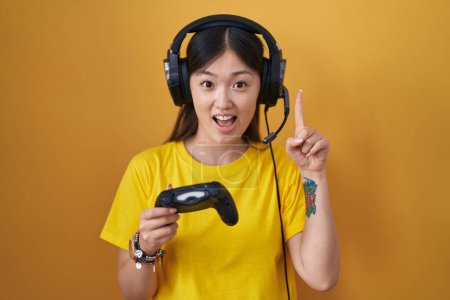 Foto de Joven china jugando videojuego sosteniendo el controlador apuntando con el dedo hacia arriba con una idea exitosa. exitado y feliz. número uno. - Imagen libre de derechos