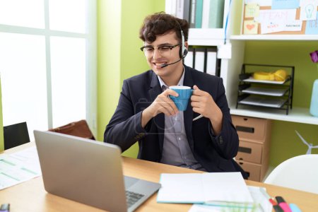 Foto de Hombre no binario agente de centro de llamadas beber café trabajando en la oficina - Imagen libre de derechos