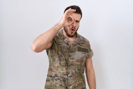 Foto de Joven hombre hispano con uniforme camuflaje del ejército haciendo un gesto bien sorprendido con la cara sorprendida, los ojos mirando a través de los dedos. expresión incrédula. - Imagen libre de derechos