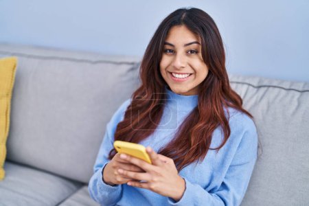 Photo for Young hispanic woman using smartphone sitting on sofa at home - Royalty Free Image