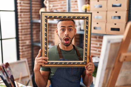 Photo for Young hispanic man sitting at art studio with empty frame afraid and shocked with surprise and amazed expression, fear and excited face. - Royalty Free Image