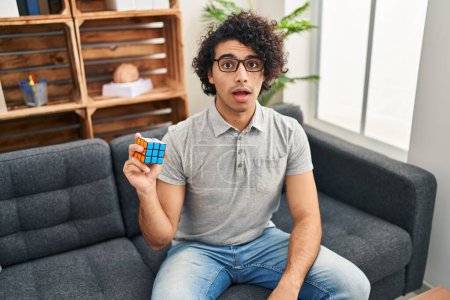 Photo for Hispanic man with curly hair playing colorful puzzle cube intelligence game scared and amazed with open mouth for surprise, disbelief face - Royalty Free Image