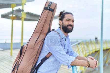 Téléchargez les photos : Jeune musicien hispanique tenant un étui de guitare appuyé sur la balustrade au bord de la mer - en image libre de droit