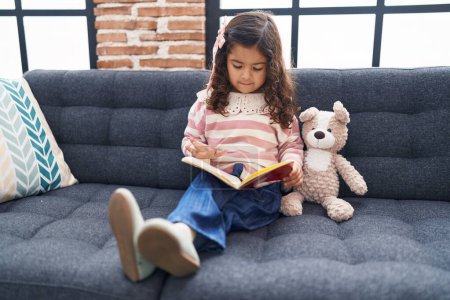 Photo for Adorable hispanic girl reading book sitting on sofa at home - Royalty Free Image