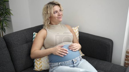 Photo for Young pregnant woman touching belly sitting on sofa at home - Royalty Free Image