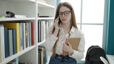 Téléchargez les photos : Jeune étudiante blonde parlant sur smartphone tenant livre à l'université salle de classe - en image libre de droit
