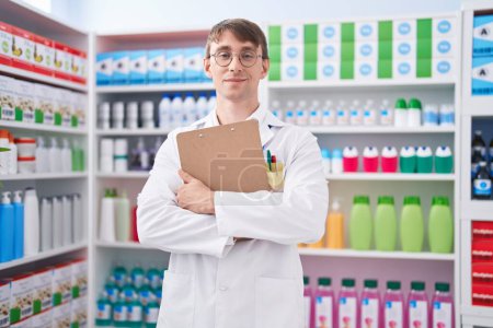Foto de Joven hombre caucásico farmacéutico sonriendo confiado sujetando portapapeles en la farmacia - Imagen libre de derechos