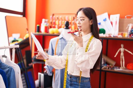 Foto de Joven mujer china sastre hablando en el teléfono inteligente que busca diseño de ropa en atelier - Imagen libre de derechos