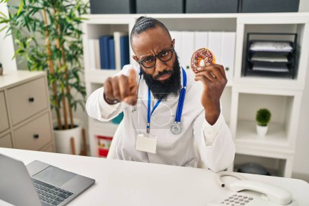 Téléchargez les photos : Afro-Américain travaillant à la clinique diététiste tenant beignet pointant du doigt la caméra et à vous, geste confiant regardant sérieux - en image libre de droit