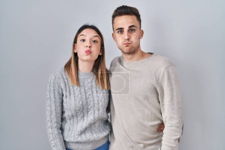 Foto de Young hispanic couple standing over white background puffing cheeks with funny face. mouth inflated with air, crazy expression. - Imagen libre de derechos
