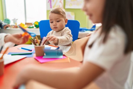 Foto de Adorable niño rubio estudiante preescolar sentado en el dibujo de la mesa en papel en el jardín de infantes - Imagen libre de derechos