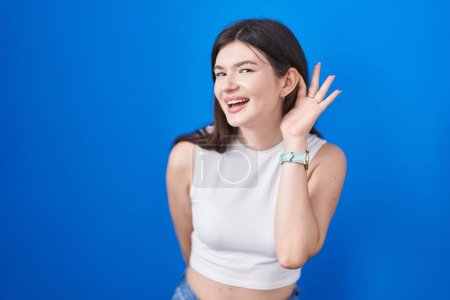 Photo for Young caucasian woman standing over blue background smiling with hand over ear listening an hearing to rumor or gossip. deafness concept. - Royalty Free Image