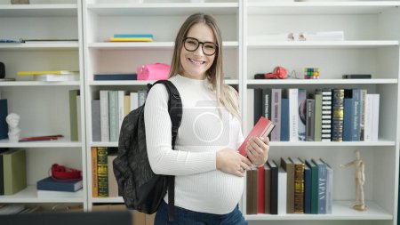 Téléchargez les photos : Jeune étudiante enceinte souriante confiante tenant des livres à la bibliothèque de l'université - en image libre de droit