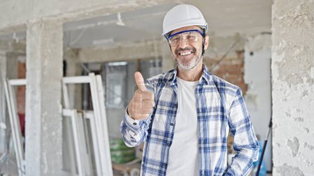 Foto de Constructor hombre de mediana edad de pie con los brazos cruzados gesto haciendo el pulgar hacia arriba gesto en el sitio de construcción - Imagen libre de derechos