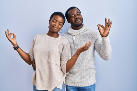 Foto de Joven pareja afroamericana de pie sobre fondo azul juntos relajados y sonriendo con los ojos cerrados haciendo gesto de meditación con los dedos. concepto de yoga. - Imagen libre de derechos