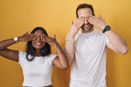 Téléchargez les photos : Couple Interracial debout sur fond jaune couvrant les yeux avec les mains souriantes gaies et drôles. concept aveugle. - en image libre de droit