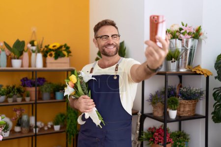 Foto de Florista hombre de mediana edad hacer selfie por teléfono inteligente con flores en la tienda de flores - Imagen libre de derechos