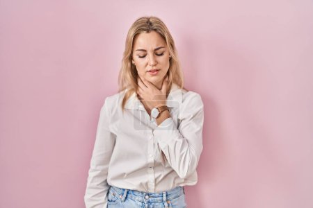 Foto de Mujer caucásica joven con camisa blanca casual sobre fondo rosa tocando el cuello doloroso, dolor de garganta para la gripe, la clod y la infección - Imagen libre de derechos