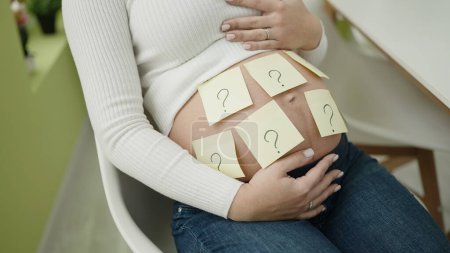 Photo for Young pregnant woman sitting on chair with question mark reminder paper on belly asking for baby gender at dinning room - Royalty Free Image