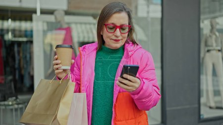 Photo for Mature hispanic woman with grey hair smiling going shopping using smartphone at clothing store - Royalty Free Image
