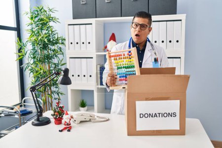 Photo for Hispanic young doctor man donating toys at the clinic in shock face, looking skeptical and sarcastic, surprised with open mouth - Royalty Free Image