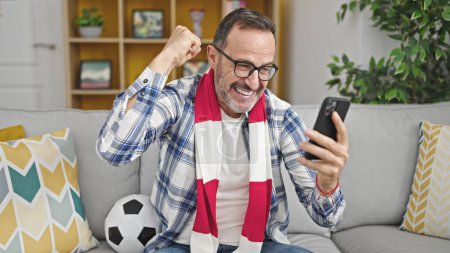 Foto de Hombre de mediana edad que apoya al equipo de fútbol viendo el juego en el teléfono inteligente en casa - Imagen libre de derechos