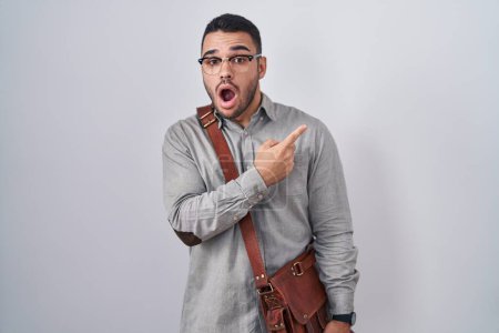 Foto de Joven hispano con maleta sorprendida señalando con el dedo hacia un lado, boca abierta expresión asombrada. - Imagen libre de derechos