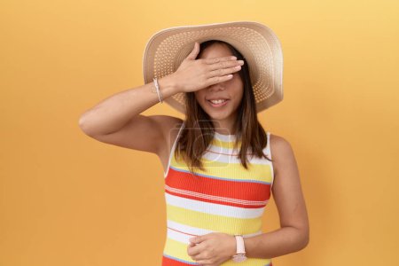 Photo for Middle age chinese woman wearing summer hat over yellow background smiling and laughing with hand on face covering eyes for surprise. blind concept. - Royalty Free Image
