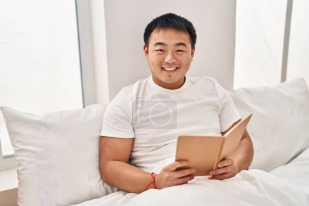 Foto de Joven hombre chino leyendo libro sentado en la cama en el dormitorio - Imagen libre de derechos
