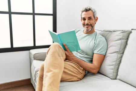 Téléchargez les photos : Moyen-âge homme aux cheveux gris livre de lecture assis sur le canapé à la maison - en image libre de droit