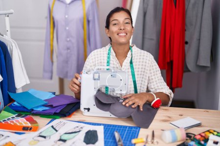 Téléchargez les photos : Jeune belle femme hispanique tailleur souriant confiant en utilisant une machine à coudre au magasin de tailleur - en image libre de droit