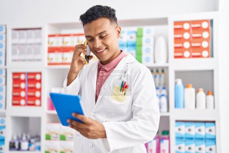 Téléchargez les photos : Jeune pharmacien homme latin parlant sur smartphone tenant bouteille de pilules à la pharmacie - en image libre de droit