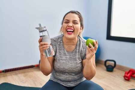 Téléchargez les photos : Jeune femme hispanique tenant pomme verte et bouteille d'eau souriant et riant fort à haute voix parce que drôle blague folle. - en image libre de droit
