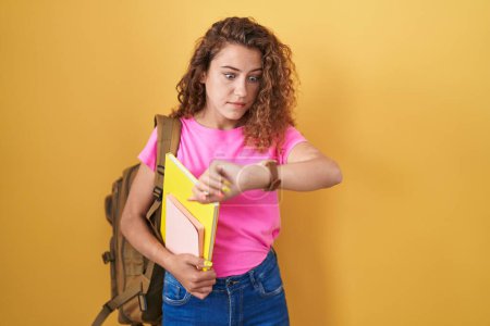Téléchargez les photos : Jeune femme caucasienne portant un sac à dos étudiant et tenant des livres regardant l'heure de la montre inquiète, peur de se retrouver en retard - en image libre de droit