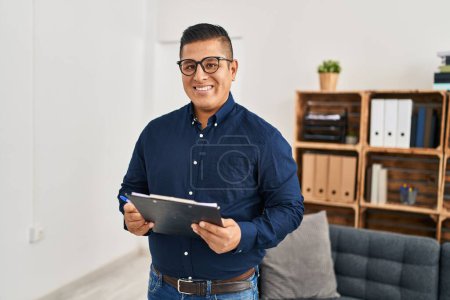 Foto de Joven trabajador de negocios latino sonriendo confiado sosteniendo portapapeles en la oficina - Imagen libre de derechos