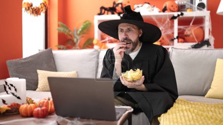 Photo for Young bald man wearing wizard costume eating chips potatoes watching movie at home - Royalty Free Image