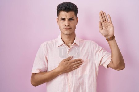 Photo for Young hispanic man standing over pink background swearing with hand on chest and open palm, making a loyalty promise oath - Royalty Free Image