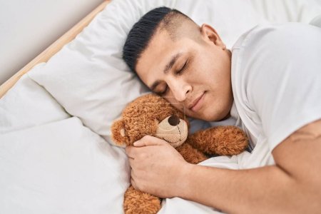 Téléchargez les photos : Jeune homme latino étreignant ours en peluche couché sur le lit dormant dans la chambre - en image libre de droit