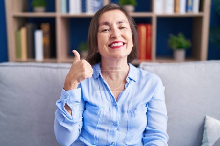 Foto de Mujer hispana de mediana edad sentada en el sofá de casa haciendo un gesto feliz con la mano. aprobando la expresión mirando a la cámara mostrando éxito. - Imagen libre de derechos