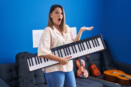 Foto de Mujer rubia sosteniendo el teclado del piano asustada y sorprendida con la boca abierta para sorpresa, cara de incredulidad - Imagen libre de derechos